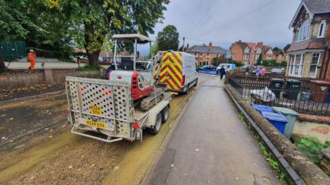 Road with homes along the right hand side - the road is covered in debris from flooding a Thames Water van and trailer with digger is parked up on the road.
