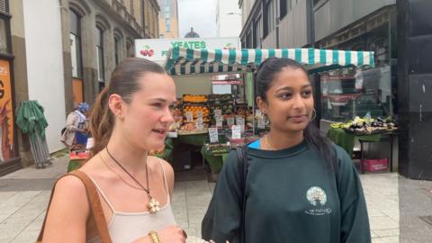 Two girls standing in front of a fruit stall on Cardiff's Queen Street. One is wearing a cream strappy top, large flower pendant and brown shoulder bag, and the other is wearing a dark green sweatshirt, large silver hoop earrings and a black shoulder bag