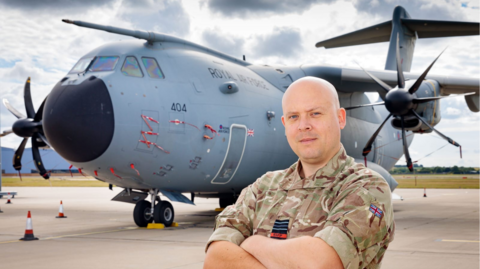 Cameron Thomas MP in military fatigues in the foreground with his arms crossed. He is stood in front of a grey Hercules plane.