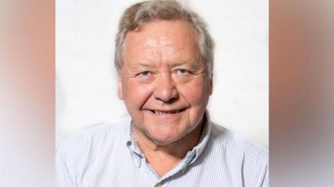 A man in a striped blue-and-white shirt with an open collar smiles against a white backdrop. He has grey hair and blue eyes.