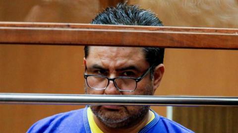 Naasón Joaquín García, wearing a yellow T-shirt below a blue overall looks over the rim of his glasses at a court appearance in Los Angeles in 2019. He has a greying beard and his black hair is cut short. 