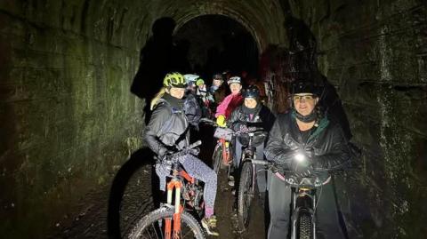 A group of woman in bikes wear helmets and warm jackets as they pose for a night-time picture in a tunnel. One of the cyclists is holding a torch.