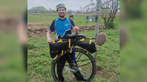 Luke Evison standing next to his unicycle wearing a blue and black t-shirt and a yellow and black cycling helmet.  He has brown hair and a brown beard and is smiling at the camera. Mr Evison is standing on grass with a stone wall and fields behind him, with two horses visible. 