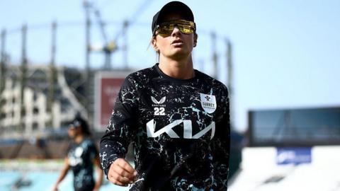 Danni Wyatt Hodge warms up ahead while playing for Surrey, dressed in a dark, patterned top, baseball cap and sunglasses at the Oval with the gas tower, stands, blue sky and a fellow player in the background