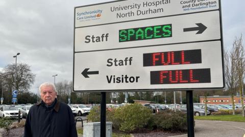 Parish councillor Grenville Holland, wearing a dark winter coat, is standing in front of a sign for the car park. A full car park is visible in the background. The sign itself if white and has digital elements. The digital parts display the words "full" in red next to an arrow to visitor and staff parking. An arrow pointing in the opposite direction labelled 'staff' says 'spaces' in green.