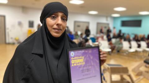 Robina Begum holds a purple and yellow award certificate from the police and crime commissioner. She is wearing black, with a head covering. The rest of the group are sitting together in the background.