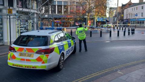 A police officer lifting up tape blocking a city centre street, next to a parked police car