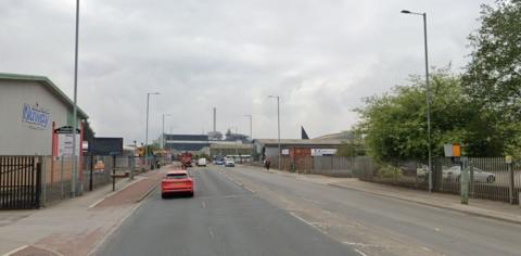 A two-lane carriageway with a red car driving southbound. There are speed cameras on both sides of the road and vehicles stopped at red traffic lights in the far distance.