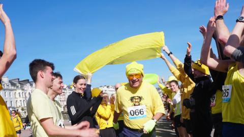 Ian Allenby walkes wearing a yellow t-shirt and hat, people hold a yellow flag above him as they cheer around him.