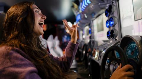 A woman with long brown hair sits an a racing car gaming machine.