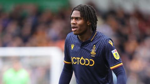 A close-up action photo of a football player wearing a dark navy Bradford City kit with yellow trim and sponsor logos. Her is on a pitch during a match, with a blurred crowd in the background.