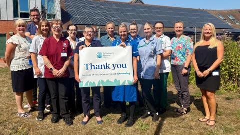 13 people posed outside a hospice building on a grassy bank in the sunshine. Several are wearing nursing uniforms. The two people at the centre are holding up a sign which says Thank You, because of you we are rated outstanding, and has the hospice logo on it.