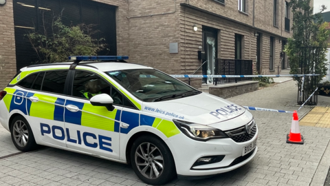 A Leicestershire Police car in front of a cordon that is in place outside a property with a smashed front door in Alexander Street, in Leicester city centre.