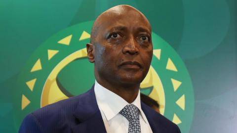 Confederation of African Football president Patrice Motsepe dressed in a suit and tie stands in front of the organisation's logo.