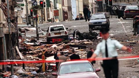 The scene of the Omagh bomb. There is a blurred police figure in front directing people away from the scene. Behind them is the wreckage from the car bomb.