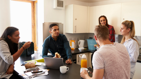 Five adults in a kitchen talking while eating.