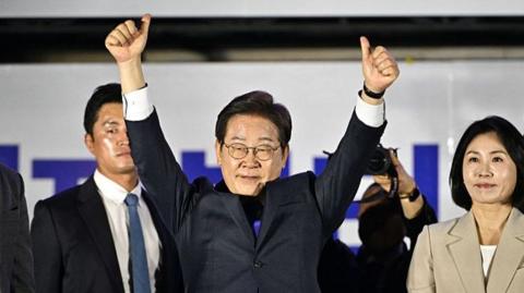 Lee Jae-myung holds two thumbs into the air as he stands next to a woman and a man, both in suits