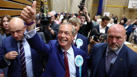 Reform UK leader Nigel Farage celebrates at the Clacton general election count on 5 July 2024, after being elected the constituency's MP. He is wearing a turquoise Reform rosette and holding his thumb up in the air. Two men wearing blue suits stand alongside him, with photographers taking pictures behind.