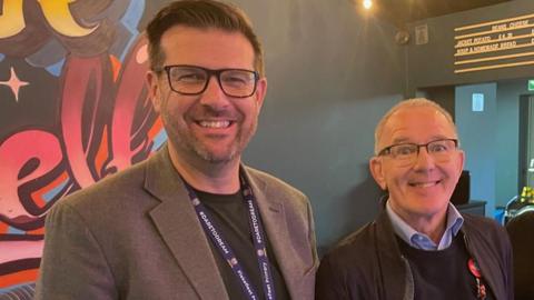 Two men - Dave McPartlin (l) and Dr Mark Spencer - are pictured standing smiling in the cafe. Both are dressed casually and wear glasses
