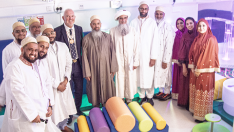 A group of people are standing in a curved line looking at the camera. There is a group of men and women, some are wearing bright colours, others are in white. They are standing in the sensory room. 