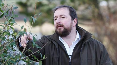 Thomas Robinson collects tea from a field. He is dressed in a white shirt, brown fleece and a brown wax jacket.