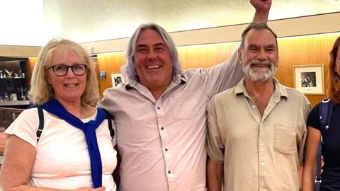 Six campaigners smile and celebrate after plans to build five affordable homes on a green space in Plymouth were rejected by councillors. They are stood inside Plymouth's Civic Centre building. The carpet is orange. One of the campaigners is holding his left arm in the air.