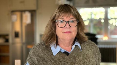 A woman with glasses and dark hair, wearing a brown knitted jumper is sitting indoors in front of a window. 