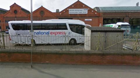 Brick-built two-storey bus depot with triangular roofed sheds and a flat-roofed garage in which a lorry is parked. A National Express coach is parked in front of the buildings.  There is a green and black railing separating the depot from the pavement.