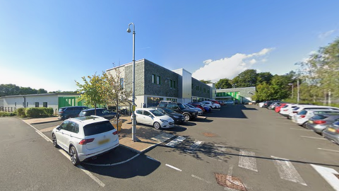 A street view image of Ferndene which is a modern, rectangular building with grey bricks on the front of the building. There are cars parked in front. Large trees can be seen behind the building.