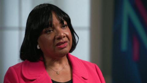 Diane Abbott, wearing a pink jacket, necklace and black top, looking at the camera during an interview