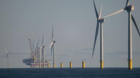 Photo of large offshore white wind turbines with the sky in the background. There are about six wind turbines in a row with a ship in the background also. The turbines are spinning. There are some clouds in the sky.