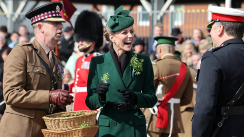 Princess of Wales wearing a bottle green coat with an Irish Guards cap star pinned to it laughing with two men in formal uniform at a St Patrick's Day parade