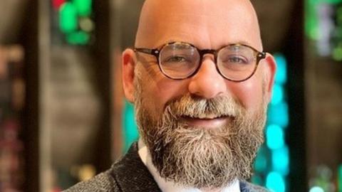 A man with a greying beard and round framed tortoiseshell glasses smiles at the camera