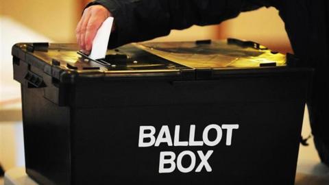 A man's hand putting a folded voting paper into a slit in a lid of a large black box with the words "Ballot Box" in white capital letters on the side.