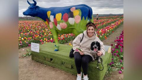 A woman sat in front of a cow sculpture in a tulip field, with her arm around a springer spaniel dog.