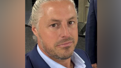 A man with greased-back blonde hair, who is wearing a suit, looks that the camera in a close-up shot