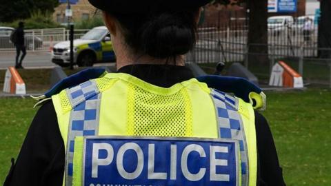A stock image of a woman police officer pictured from the back, with a tight crop showing the lower part of her heard and the top of her back. She has dark hair in a low bun. She is wearing a dark uniform with a yellow high-viz vest with the work Police on it. She is in a public place that looks like a patch of grass by a main road, where a police car is parked in the distance.