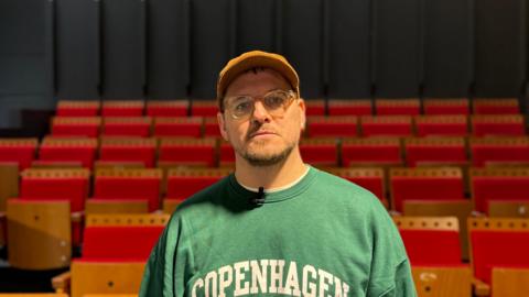 A head and shoulders shot of Paul Smith looking at the camera. He is wearing glasses, an orange cap and a green sweater with "Copenhagen" in white bold capital letters printed on the front. He is stood in front of red theatre chairs. The walls are black.