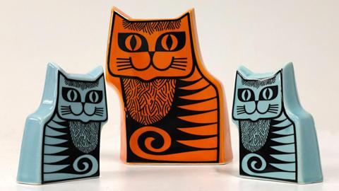 Three cat-shaped money boxes on a shelf. The middle one is the tallest and is orange, with two light blue ones either side.