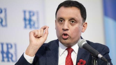 Anas Sarwar, who has short black hair, speaks into a microphone with his right hand raised next to his side. He is wearing a navy suit, white shirt and red tie.