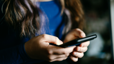 A close up stock image of an anonymous girl with long brown hair and wearing a blue top, holding a black smart phone in front of her.
