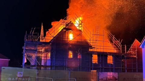 A street view of a house with scaffolding outside with orange flames and plumes of orange smoke coming out of it.