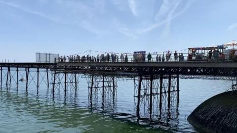 Queen's pier in Ramsey, which is a Victorian iron pier stretching out over the water in Ramsey Bay. There are crowds of people standing on it with bunting along the railings.