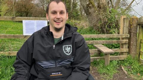 A man in a wheelchair in beside an wooden fence stile along the green trail