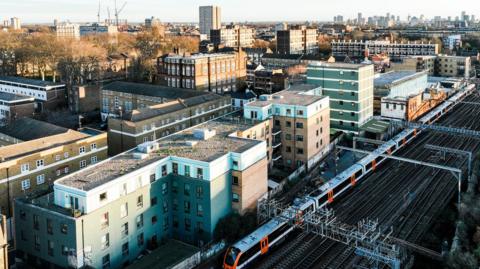 A train line runs past a residential area in London, with the London skyline in the background. The residential area mostly consists of low-rise blocks of flats. A train is running on the train tracks past the flats.