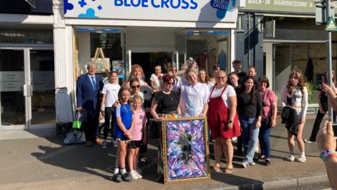 A crowd of people standing outside a Blue Cross charity shop smiling at the camera. At the front is a blonde woman in a white t-shirt holding up a mirror, painted with purple and blue patterns, in a gold frame.