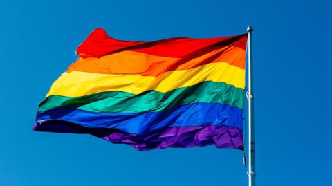 A rainbow flag is seen attached to a flag pole bearing the colours of purple, blue, green, yellow, orange and red. The flag is the universal symbol of LGBTQ pride.