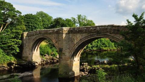 The 40ft (12m)  stone bridge consists of two support arches spanning a river. Lush, green trees border each side.