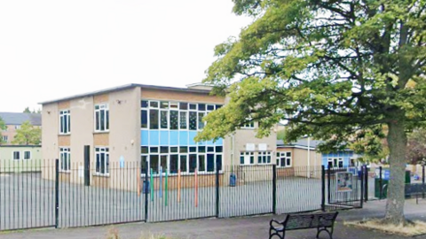 A brown square building with lots of rectangular windows behind a tall black gate. A street with a black metal bench and tall tree can also be seen