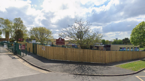 Trinity CE Primary Academy stands on the corner of a street with a wooden fence surrounding it and the entrance on the left of the photo.